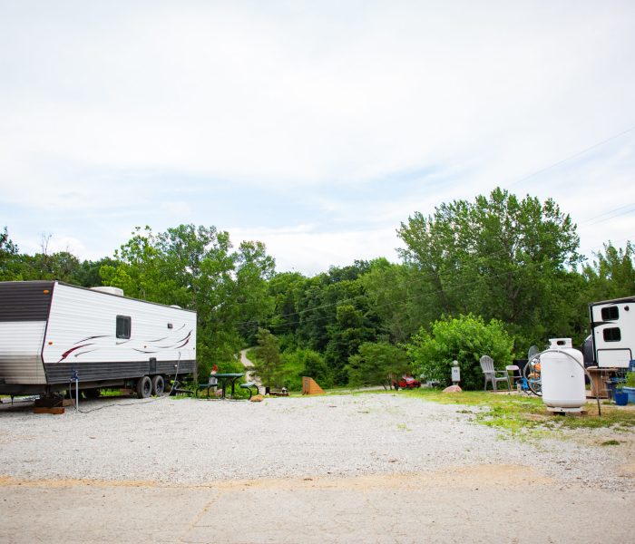 Two RVs are parked on a gravel lot surrounded by trees and greenery, perfect for a long-term stay. Outdoor chairs, a grill, and supplies are scattered around the area under a partly cloudy sky.