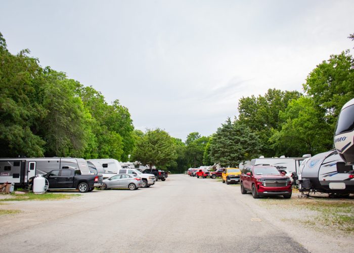 A paved road in an RV park is lined with parked RVs and vehicles on both sides, surrounded by green trees under a cloudy sky.