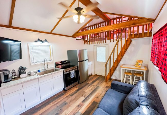 Small kitchen and living area with white cabinets, stainless steel appliances, a gray sofa, TV, and a wooden loft accessed by stairs.