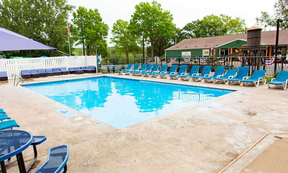 Outdoor swimming pool surrounded by blue lounge chairs, a shaded seating area, and a green building decorated with patriotic banners in the background, with trees and a fence enclosing the area.