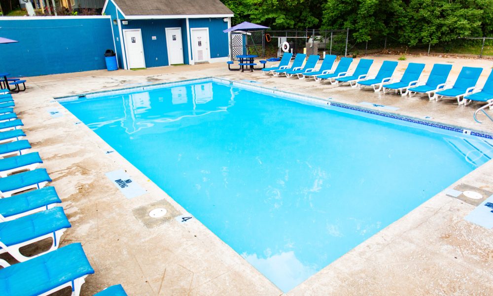 An outdoor swimming pool surrounded by blue and white lounge chairs, with a blue building and changing rooms in the background, and green trees lining the area. The pool deck is wet and empty.
