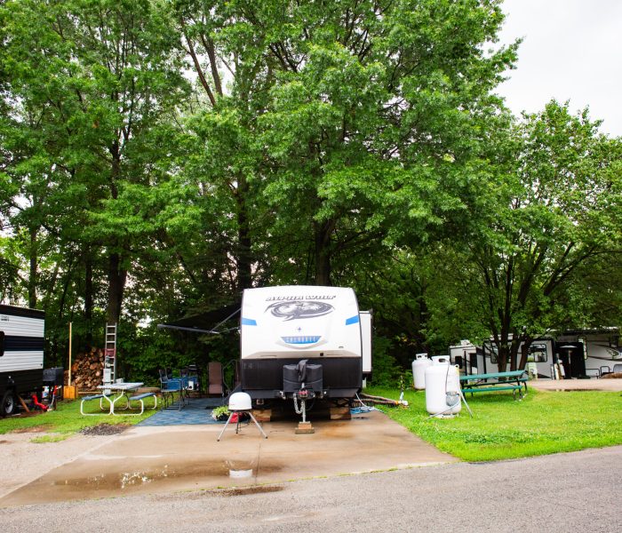 A campsite with a travel trailer parked under large green trees. Nearby are picnic tables, a barbecue grill, propane tanks, and camping chairs. The ground is wet, suggesting recent rain. Other RVs are visible in the background.