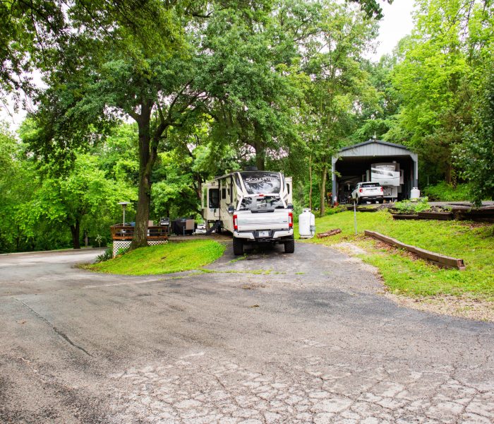 A white RV is parked on a driveway surrounded by trees and greenery. A garage with another vehicle inside is visible to the right. The area appears to be a quiet, shaded neighborhood or campground.