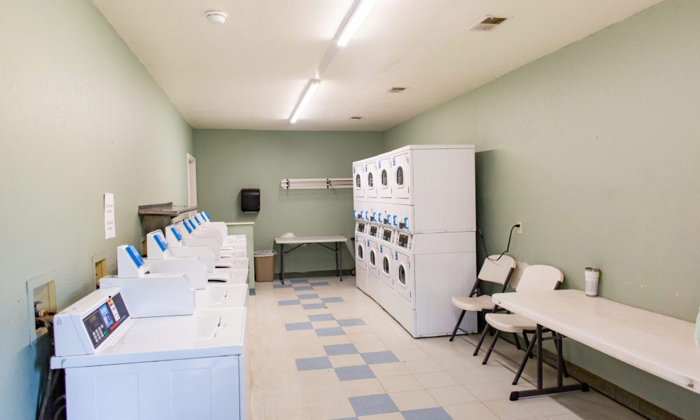 A clean laundry room with multiple white washers and dryers along two walls, folding tables, plastic chairs, pale green walls, and light blue and white tiled flooring under fluorescent lights.