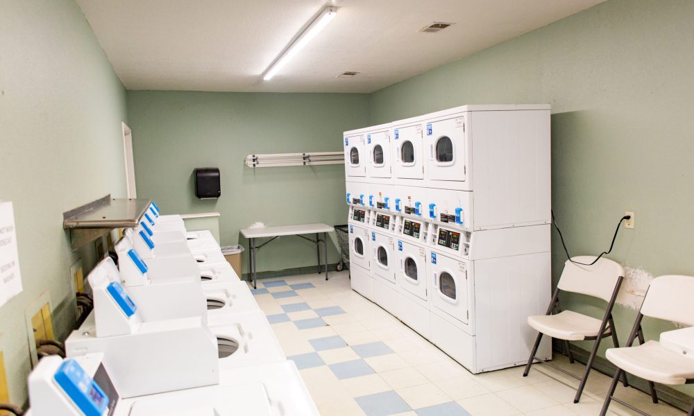 A clean, empty laundromat room with washing machines on the left, stacked dryers on the right, plastic chairs, a folding table, and pale green walls with fluorescent ceiling lighting.