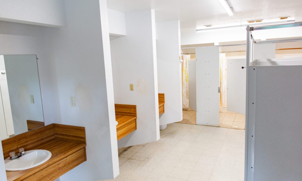 A clean, white restroom with wooden countertops, sinks, mirrors, and partially open shower stalls with curtains in the background. The floor is tiled, and fluorescent lights are on the ceiling.