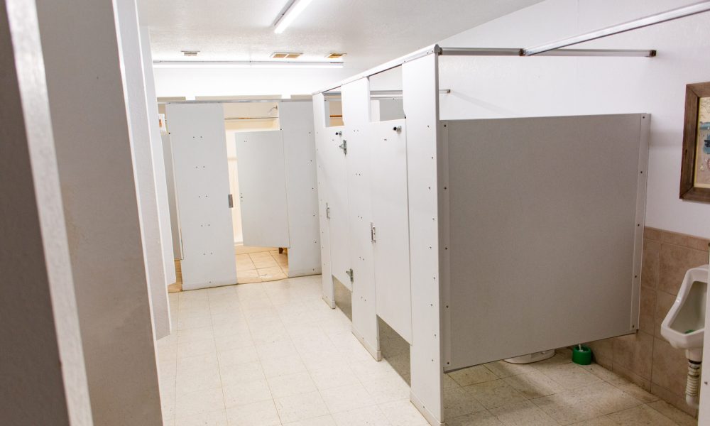 A clean public restroom with multiple white stalls, beige tiled walls, and a urinal visible on the right side; fluorescent ceiling lights illuminate the space.