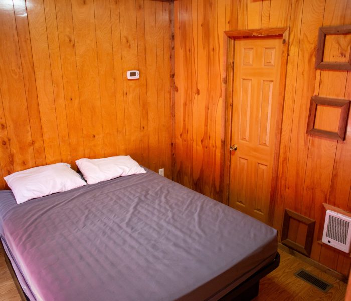 A cozy, wood-paneled bedroom ideal for a long-term stay, featuring a queen bed with gray sheets and white pillows. The room includes a wooden door, empty picture frames on the wall, and a thermostat above the light switch.