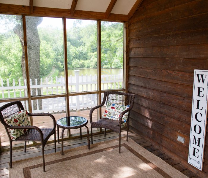 A cozy screened porch with two wicker chairs, a glass-top table, and a welcome sign on a wooden wall. Perfect for a long-term stay, the large windows reveal green trees and a white picket fence outside.
